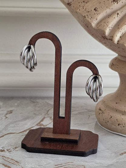 Tazmina Hoop Earrings in Silver featuring a polished, multi-layered curved hoop design with a shiny silver finish, displayed on a dark wooden jewellery stand.