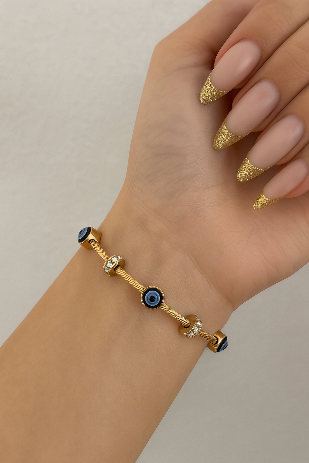 Gold cable bangle bracelet with blue evil eye charms and cubic zirconia spacers displayed on a neutral cushion.
