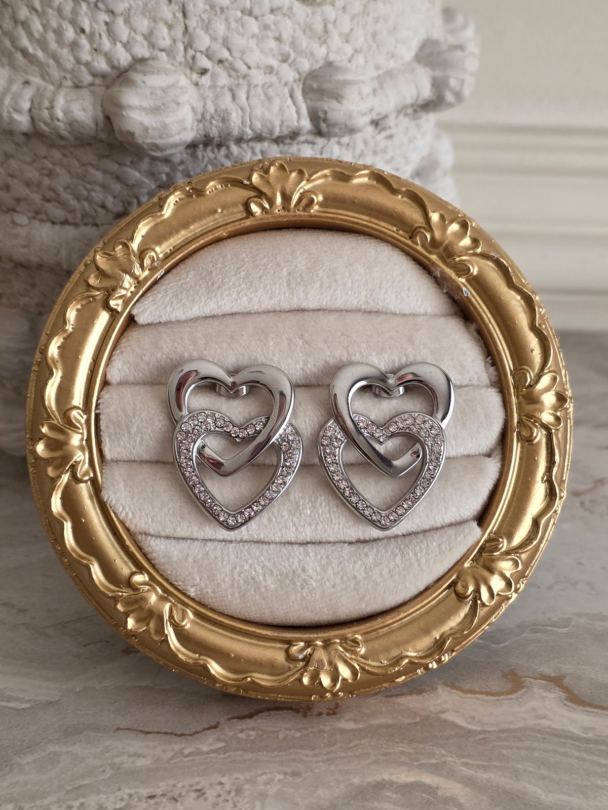 Liana Heart Earrings Silver featuring intertwined polished silver-tone hearts, one adorned with sparkling clear rhinestones, displayed on a cream velvet jewellery stand inside an ornate gold frame.