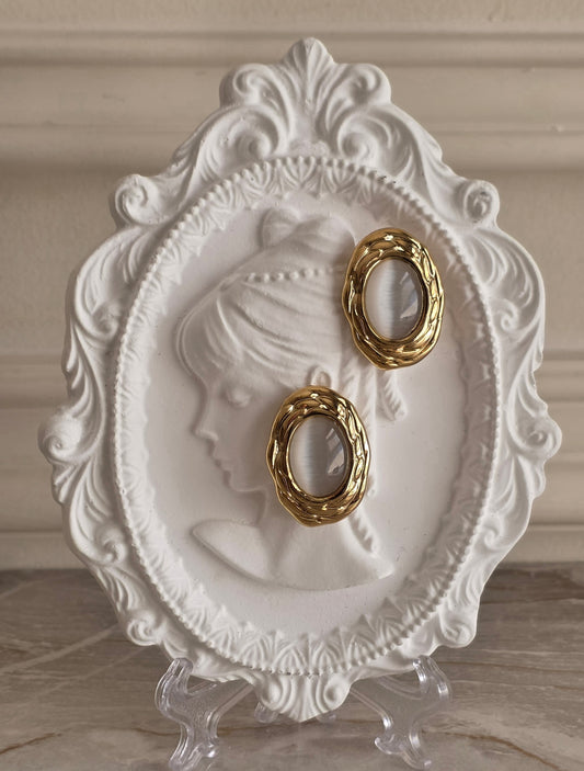 Maya Earrings featuring oval grey cat-eye stones framed in textured gold, displayed on an elegant white vintage-style jewellery plate.